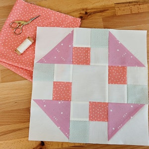 Puede incluir: Un bloque de patchwork con un cuadrado blanco central, rodeado de piezas de tela estampadas en rosa, blanco y azul claro. Un trozo de tela rosa con lunares blancos, unas tijeras doradas y una bobina de hilo están junto al bloque. Suministros de costura.