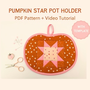 Può includere: Un sottopentola a forma di zucca con un design a stella, con uno sfondo marrone con stelle bianche e una stella rosa al centro. Il sottopentola ha un bordo rosa e un anello per appenderlo. Il testo "PUMPKIN STAR POT HOLDER" è in alto.