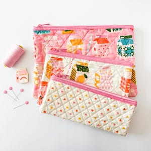 May include: Three quilted zipper pouches in varying sizes, featuring pink and white fabrics with colorful milk carton and heart patterns. A spool of pink thread, pins, and a milk carton pin are also present.