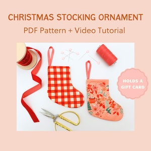 Pode incluir: Dois enfeites de meia de Natal, um xadrez vermelho e branco, o outro com um padrão floral. A imagem inclui fita vermelha, tesoura, linha e alfinetes. Texto: Enfeite de meia de Natal, padrão PDF + tutorial em vídeo. Contém um cartão-presente.