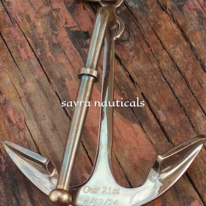 May include: A bronze anchor with the inscription "Our 21st 4/12/24". The anchor is displayed against a rustic, weathered wooden background. The design features a ring, a shank, and two flukes. The text "sayra nauticals" is also visible.