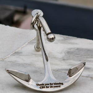 May include: A shiny silver anchor with a curved design, set on a marble surface. The anchor has a ring and a ball at the top. The text engraved on the anchor reads: "Thank you for blessing CBS for 20 years!"