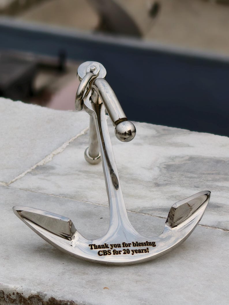 May include: A shiny silver anchor with a curved design, set on a marble surface. The anchor has a ring and a ball at the top. The text engraved on the anchor reads: "Thank you for blessing CBS for 20 years!"