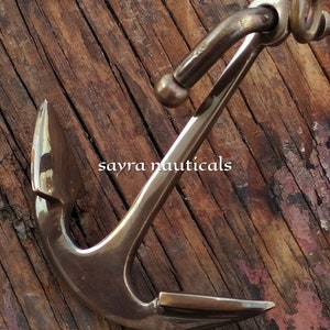 May include: A bronze anchor with a curved design, set against a weathered wooden background. The anchor features a detailed construction with a ring and a pointed fluke. The text "sayra nauticals" is visible on the image.