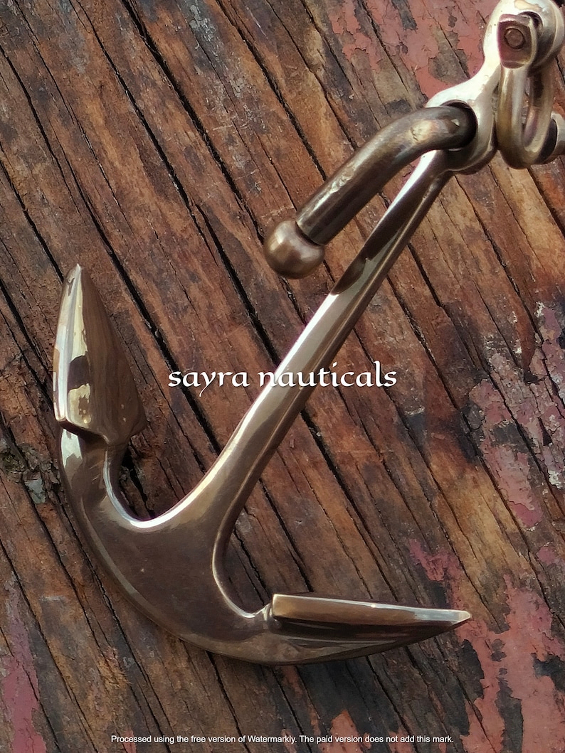 May include: A bronze anchor with a curved design, set against a weathered wooden background. The anchor features a detailed construction with a ring and a pointed fluke. The text "sayra nauticals" is visible on the image.