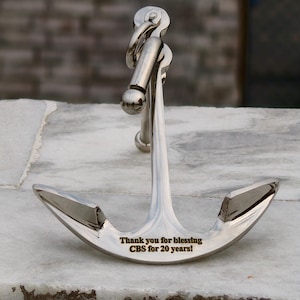 May include: A polished silver anchor with a curved design. The anchor has a ring at the top and the text "Thank you for blessing CBS for 20 years!" engraved on the front. The anchor is displayed on a white marble surface.