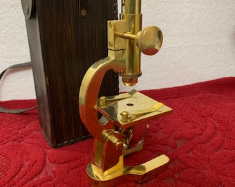 Könnte beinhalten: Ein antikes Messingmikroskop mit einer Holztasche. Das Mikroskop hat ein einzelnes Objektiv und eine Bühne zum Betrachten von Präparaten. Die Tasche ist braun mit einem Lederriemen.