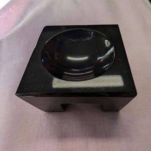 May include: A black square pedestal with a round, recessed bowl on top. The pedestal has four legs.