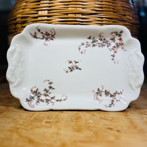 May include: A rectangular white ceramic serving tray with a scalloped edge. The tray is decorated with delicate floral patterns in blue, green, and rust colors. The tray has two handles on either side.