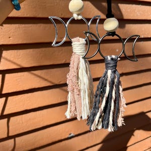 May include: Two triple moon charm car air fresheners with wooden beads and fabric tassels. One tassel is pink and white, the other is black and white.