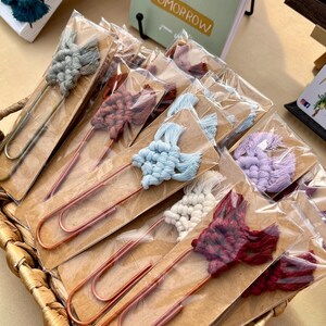 May include: Assortment of colorful macrame bookmarks with gold and copper paper clips. The bookmarks are packaged individually on brown cardstock. The text "Hurry to Tomorrow" is visible in the background.