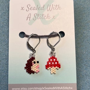 Puede incluir: Un par de pendientes de champiñón de cuentas con ganchos plateados. El champiñón de la izquierda es rojo con puntos blancos y el champiñón de la derecha es marrón con una cara rosa. Los pendientes están en una tarjeta blanca con el texto "x Sealed With A Stitch x" y la dirección del sitio web "www.etsy.com/shop/xSealedWithAStitchx" y el nombre de usuario de Instagram "@xSealedWithAStitchx".