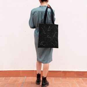 Puede incluir: Una bolsa de tela negra con diseños de constelaciones blancas. La bolsa tiene asas negras y la lleva una persona.