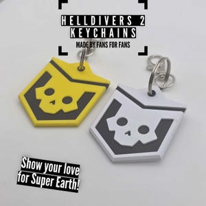 Op de afbeelding: Twee sleutelhangers met een schildontwerp met een schedel, één geel en één wit. De tekst "HELLDIVERS 2 KEYCHAINS" en "MADE BY FANS FOR FANS" zijn zichtbaar. De zin "Show your love for Super Earth!" is ook aanwezig.