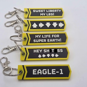 Pode incluir: Vários chaveiros amarelos e pretos com fechos prateados. Cada chaveiro tem uma inscrição diferente em texto branco. As frases incluem "SWEET LIBERTY MY LEG!", "MY LIFE FOR SUPER EARTH!", "HEY SH TSS" e "EAGLE-1".