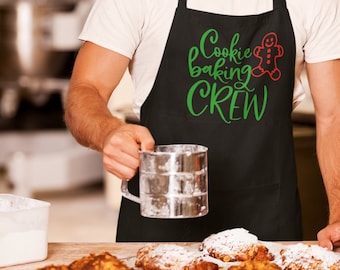 Może przedstawiać: Czarny fartuch z napisem "Cookie baking CREW" w kolorze zielonym i czerwoną grafiką piernikowego ludzika. Osoba trzyma metalowe sito. Wypieki znajdują się na tacy na pierwszym planie.