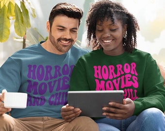 Może przedstawiać: Mężczyzna i kobieta siedzą na brązowej sofie, oboje w bluzach z napisem "HORROR MOVIES" w stylu retro. Mężczyzna trzyma biały kubek, a kobieta tablet. Bluzy są w kolorze morskim i ciemnozielonym.