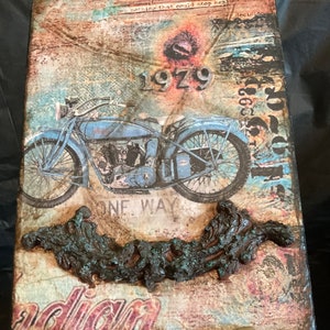 May include: Mixed media artwork featuring a vintage blue motorcycle with the year "1979" in silver. The artwork has a distressed, textured background with the word "Indian" at the bottom. A textured, dark brown sculptural element is at the bottom.