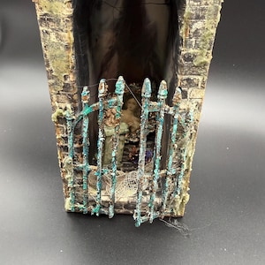 May include: A miniature diorama featuring a tall, arched structure with a weathered brick texture and a rusted, teal-colored metal gate. Inside the enclosure, a small figurine stands behind the gate, surrounded by textured netting and debris.
