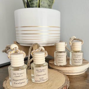 May include: Four small glass car air freshener bottles with wooden caps and hanging ropes. The labels read MG Designs & Candle Co. with scents Peppermint & Eucalyptus, Clean Cotton, Mystic Waters, and Warm Apple Bourbon. Each bottle contains clear liquid.