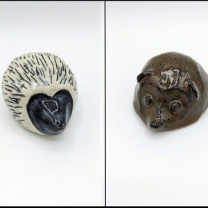 May include: Two ceramic hedgehog figurines. One is cream-colored with black markings and a black face. The other is brown with a glossy finish. Both are small and appear handmade, showcasing a whimsical animal theme.