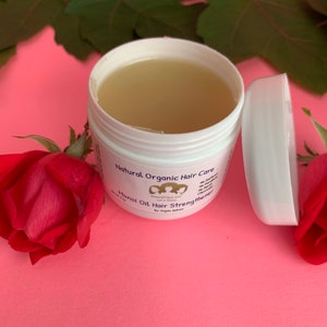 May include: A white jar with a light yellow liquid inside. The jar is labeled "Natural Organic Hair Care" and "Honoi Oil Hair Strengthener" with a purple logo. The jar is sitting on a pink surface with a red rose and green leaves.