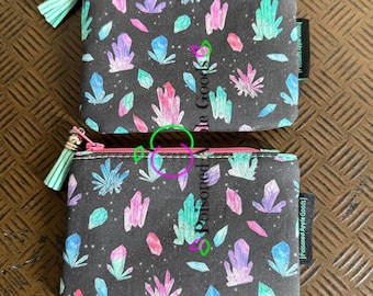 Peut inclure: Deux petites pochettes rectangulaires à fermeture éclair, avec un fond noir et un motif de cristaux colorés. Les pochettes ont une bordure rose, des pompons vert menthe et le texte "Poisoned Apple Goods".