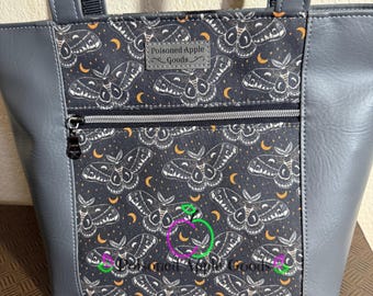 Pode incluir: Bolsa tote cinza com um bolso com zíper. A bolsa apresenta um tecido cinza escuro com um padrão repetido de mariposas e luas crescentes. O texto "Poisoned Apple Goods" é visível em uma pequena etiqueta.