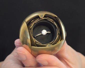 May include: A gold-colored, spherical ring box, open to reveal a diamond engagement ring. The ring has a large, round center stone and a band with smaller diamonds. The box is held open against a black background.