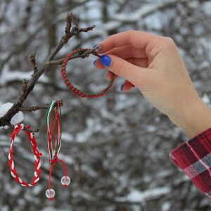 Può includere: Tre braccialetti dell'amicizia colorati appesi a un ramo d'albero spoglio in un paesaggio invernale innevato. I braccialetti sono rosso, bianco e verde, e uno ha delle perline bianche.