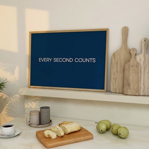 May include: A blue framed sign with the text "EVERY SECOND COUNTS" hanging on a white wall above a kitchen counter. The counter has a wooden chopping board with sliced bread, two ceramic mugs, and a cup of coffee. There are also two wooden chopping boards and a few green pears on the counter.