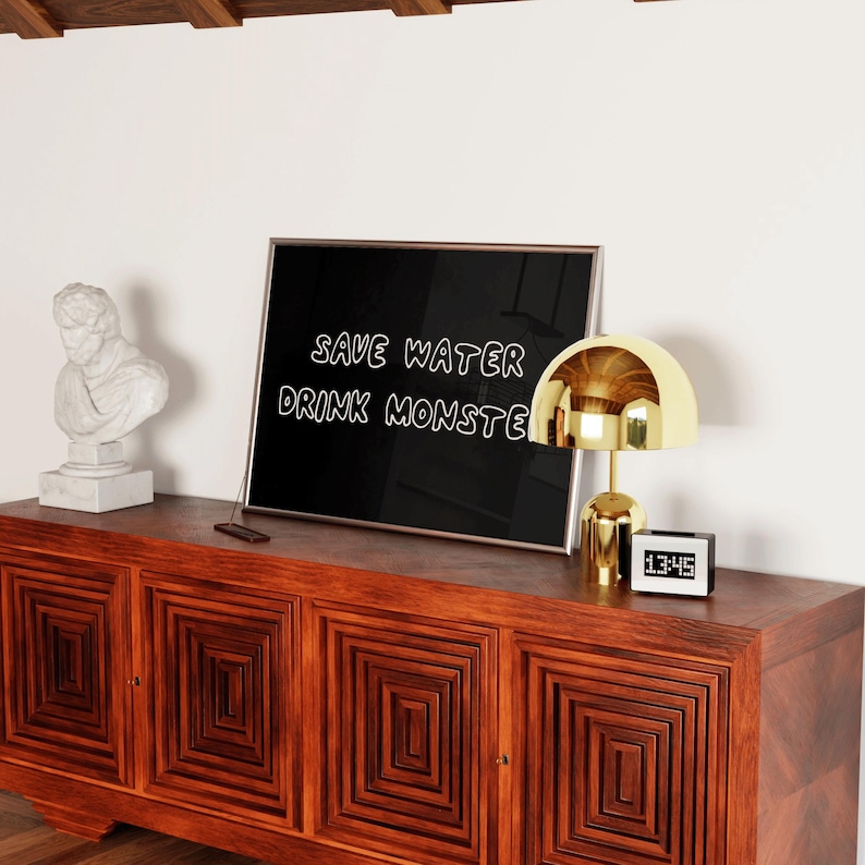 May include: A wooden cabinet with a geometric design, topped with a framed black sign that reads "SAVE WATER DRINK MONSTE". A gold-colored lamp and a digital clock are on the cabinet. A white bust sculpture is on the left.