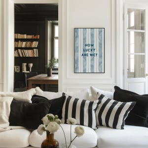 May include: A white couch with black and white striped throw pillows in a living room. A framed print with blue watercolor stripes and the text "How lucky are we?" hangs on the wall.