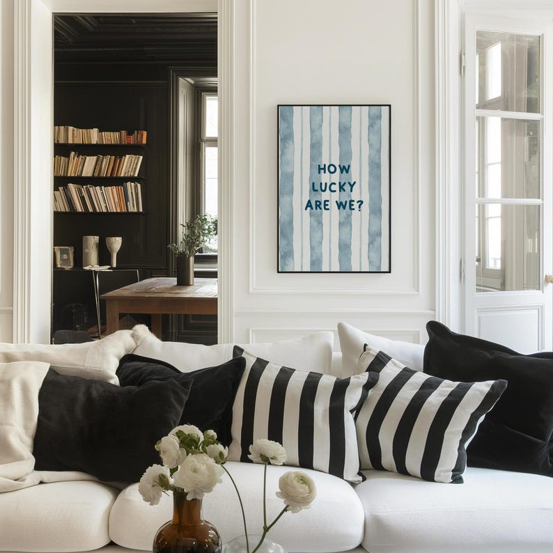 May include: A white couch with black and white striped throw pillows in a living room. A framed print with blue watercolor stripes and the text "How lucky are we?" hangs on the wall.