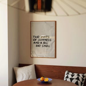 May include: A framed poster with the text "TWO PINTS OF GUINNESS AND A BIG FAT SNOG" hangs on a wall. The poster is in a room with a wooden table, a blue plate with oranges, and a white pillow.