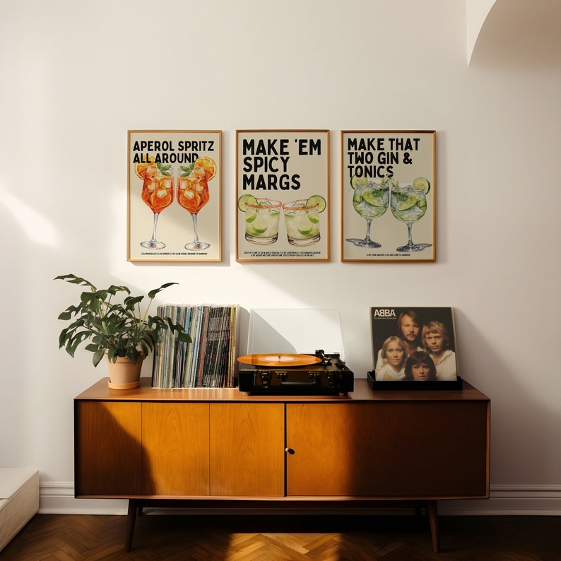 May include: Three framed prints with illustrations of cocktails and text. The prints are hung on a white wall above a wooden cabinet with a record player and a record on top. The record is by ABBA.