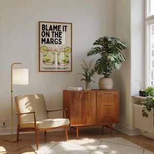 May include: A room interior featuring a framed poster with the text "BLAME IT ON THE MARGS" above a cocktail illustration. The room includes a mid-century modern style wooden cabinet, a floor lamp, a beige armchair, and a large potted plant.