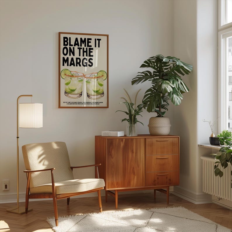 May include: A room interior featuring a framed poster with the text "BLAME IT ON THE MARGS" above a cocktail illustration. The room includes a mid-century modern style wooden cabinet, a floor lamp, a beige armchair, and a large potted plant.