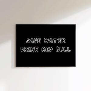 May include: A framed black print with white text that reads "SAVE WATER DRINK RED BULL". The text is in a handwritten style. The print is displayed on a white wall.