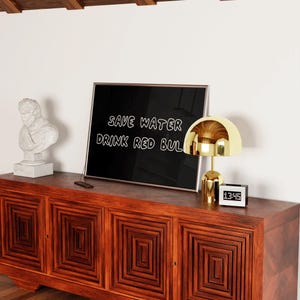 May include: A wooden cabinet with a geometric design holds a framed sign that reads "SAVE WATER DRINK RED BUL". A gold-colored lamp and a digital clock are on the cabinet. A white bust statue is on the left.