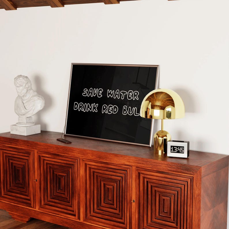 May include: A wooden cabinet with a geometric design holds a framed sign that reads "SAVE WATER DRINK RED BUL". A gold-colored lamp and a digital clock are on the cabinet. A white bust statue is on the left.