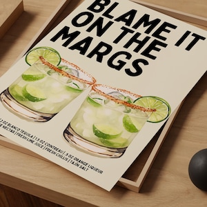 May include: A print featuring two margarita cocktails with lime wedges and a Tajin salt rim. The text "BLAME IT ON THE MARGS" is displayed in bold black letters. The ingredients are listed below the cocktails.
