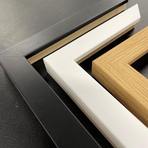 May include: Close-up of three picture frame corners in black, white, and natural wood tones. The frames are angled, showcasing their construction and color variations. The black frame is on the left, followed by a white frame, and a natural wood frame.