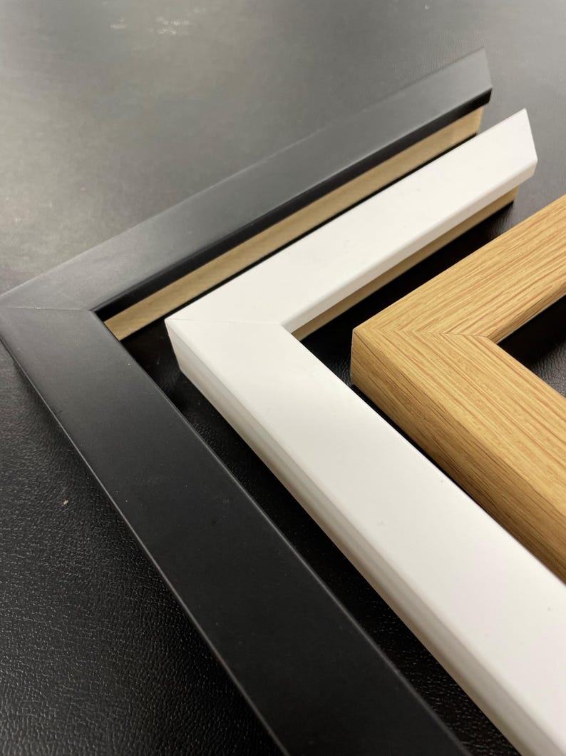 May include: Close-up of three picture frame corners in black, white, and natural wood tones. The frames are angled, showcasing their construction and color variations. The black frame is on the left, followed by a white frame, and a natural wood frame.