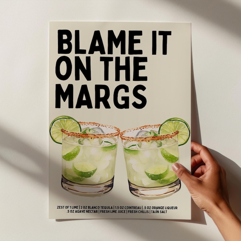 May include: A print featuring two margarita cocktails with lime wedges and a salt-rimmed glass. The text "BLAME IT ON THE MARGS" is displayed above the cocktails. The ingredients are listed below the cocktails.
