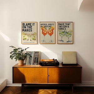 May include: Three framed art prints with cocktail illustrations hang above a wooden cabinet. The prints read "BLAME IT ON THE MARGS", "APEROL SPRITZ ALL AROUND", and "MAKE THAT TWO GIN & TONICS". A record player and plant are on the cabinet.