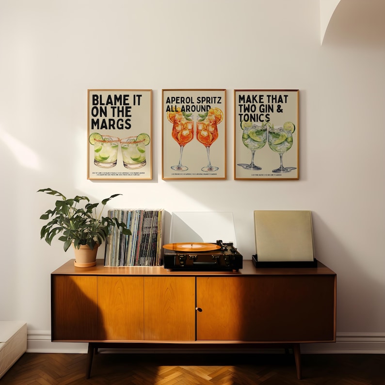 May include: Three framed art prints with cocktail illustrations hang above a wooden cabinet. The prints read "BLAME IT ON THE MARGS", "APEROL SPRITZ ALL AROUND", and "MAKE THAT TWO GIN & TONICS". A record player and plant are on the cabinet.