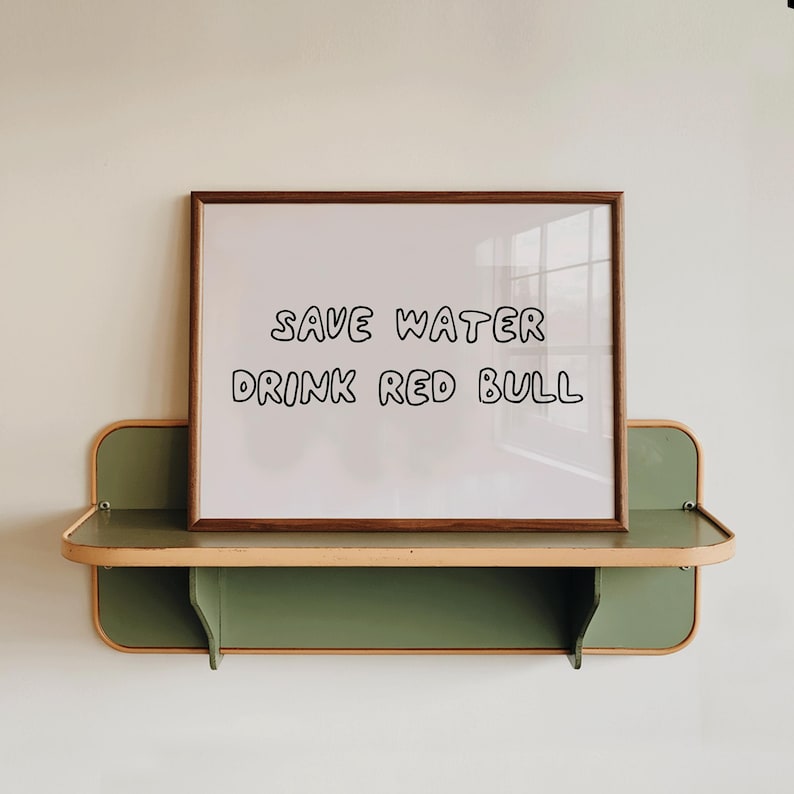 May include: A framed white sign with the text "SAVE WATER DRINK RED BULL" sits on a green and tan shelf. The frame is a dark brown color. The shelf is mounted on a white wall.