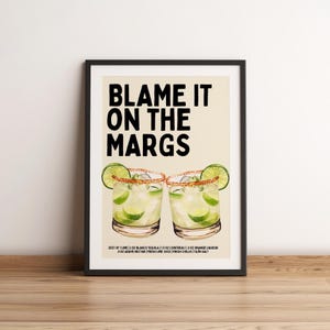 May include: A framed print with the text "BLAME IT ON THE MARGS" above two margarita cocktails. The cocktails are garnished with lime wedges and salt rims. The ingredients are listed below the cocktails.