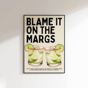 May include: A framed art print with the text "BLAME IT ON THE MARGS" in bold black letters. Below the text are two margarita cocktails with lime wedges and a recipe.
